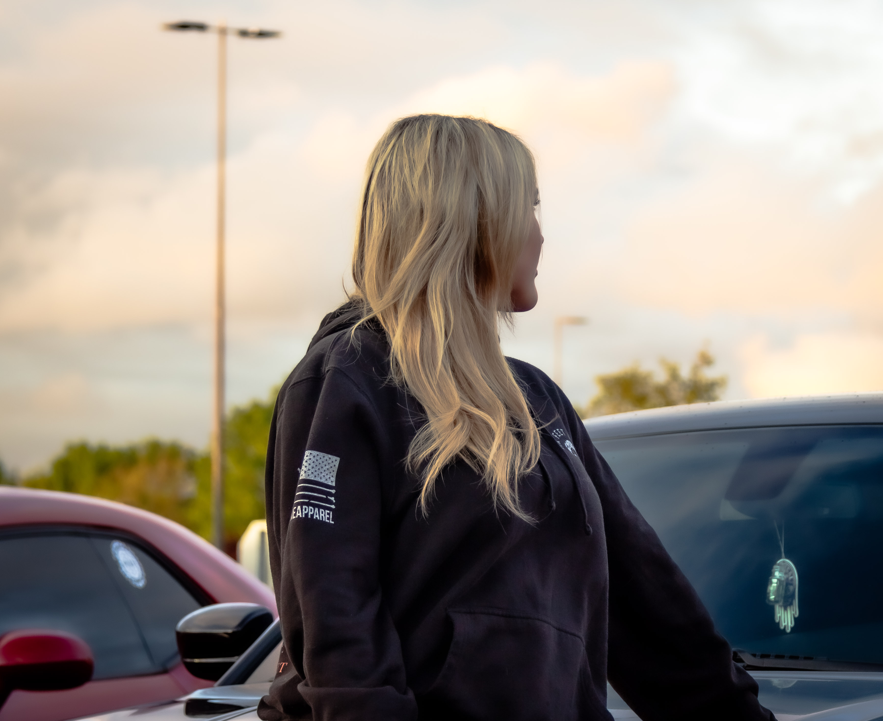Blonde woman in a black hoodie standing at a car meet during golden hour with cars visible in the background