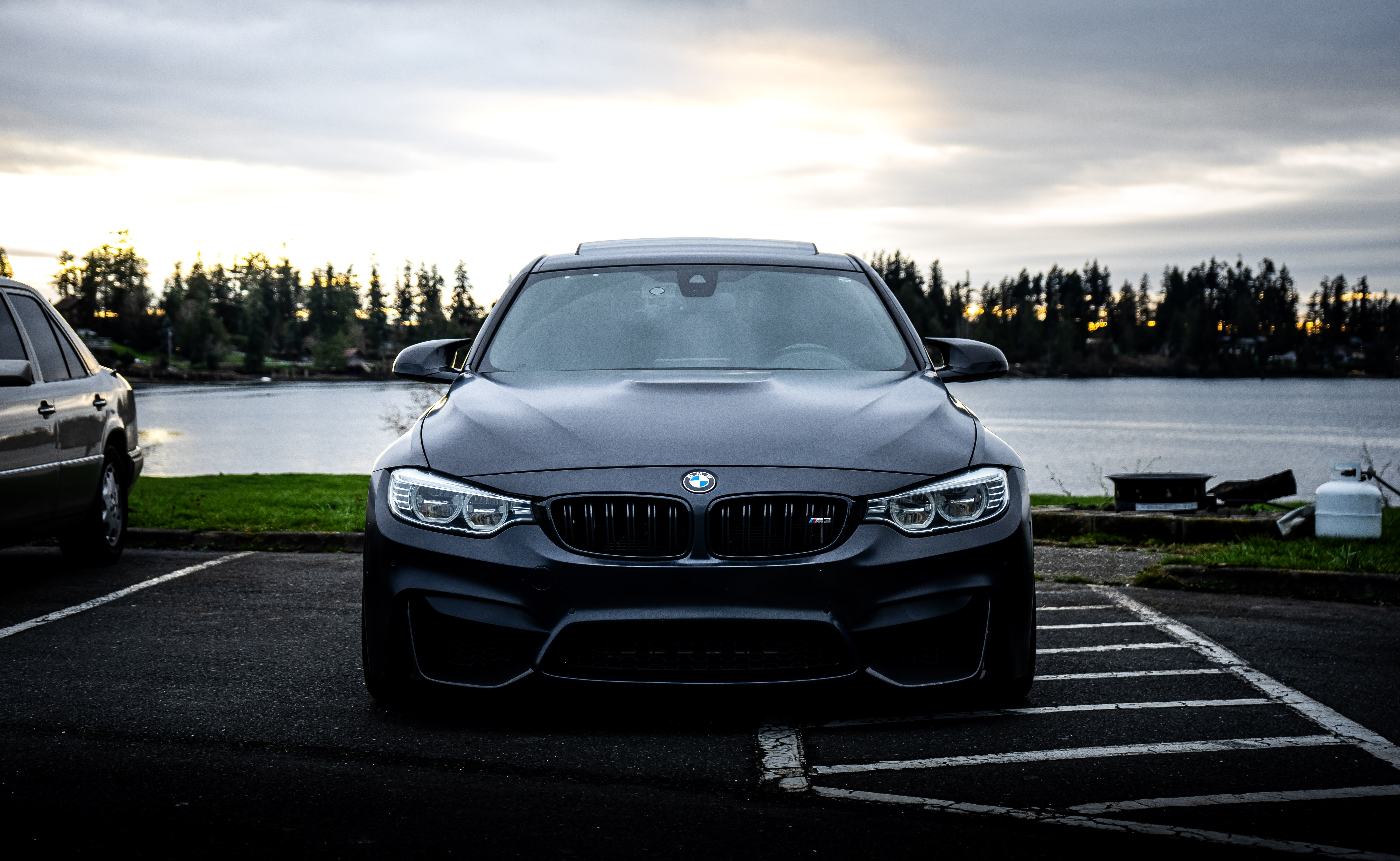Matte black BMW M3 shot front-on with a lake and dramatic overcast sky in the background